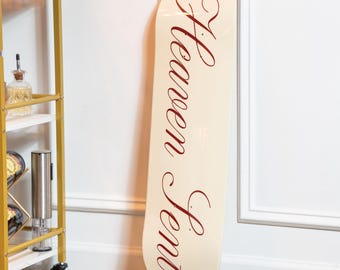 May include: Off-white skateboard with the words "Heaven Sent" in a burgundy script. The skateboard is leaning against a white wall with a decorative frame. A gold bar cart with bottles and glasses is in the background.