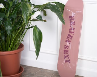 May include: A dusty rose skateboard deck featuring the text HEAVEN SENT printed in a dark purple, wavy, bold font running vertically down the center of the board. The deck has a smooth, glossy finish and a rounded nose and tail shape.