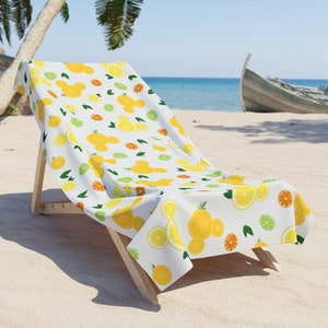 Puede incluir: Una silla de playa blanca con una toalla de playa con estampado de lim&oacute;n y lima. La toalla tiene un fondo blanco con limones amarillos, limas verdes y hojas verdes.
