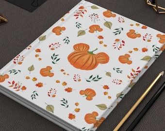 May include: A white notebook with a fall-themed design featuring orange pumpkins in the shape of Mickey Mouse ears. The pumpkins are surrounded by green leaves and orange and brown leaves. The notebook is lying on a brown surface with two pencils and a gold paperclip.
