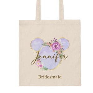 May include: A white canvas tote bag with a purple and gold floral design. The design features a stylized Mickey Mouse head with a floral wreath. The text "Jennifer Bridesmaid" is printed below the design.