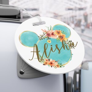 May include: A round luggage tag with a white background and a blue Mickey Mouse head with a gold outline. The Mickey Mouse head is decorated with a floral wreath in shades of orange, yellow, and pink. The name "Alisha" is written in gold cursive lettering.