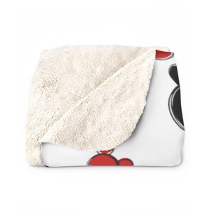 K&ouml;nnte beinhalten: Eine wei&szlig;e Fleece-Decke mit einem Design aus einem schwarzen und roten Comic-Charakter. Der Charakter hat gro&szlig;e, runde Ohren und ein breites L&auml;cheln.