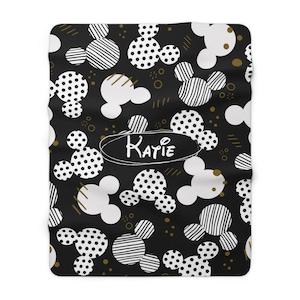 Puede incluir: Manta de forro polar de color negro y blanco con un &oacute;valo dorado que contiene el nombre "Katie" escrito a mano.