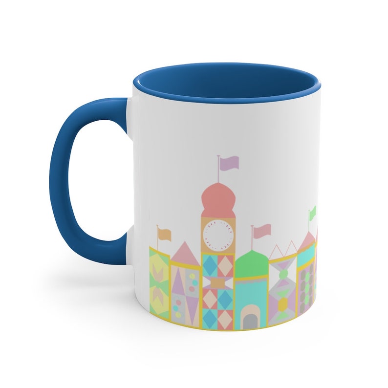 May include: A white ceramic coffee mug with a blue rim and handle. The mug features a colourful illustration of a whimsical castle with a clock tower and flags.