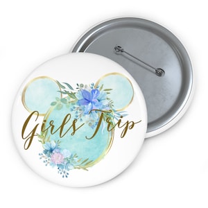 May include: A white button with a gold outline of Mickey Mouse ears and blue watercolor flowers. The text "Girls Trip" is written in gold cursive lettering.