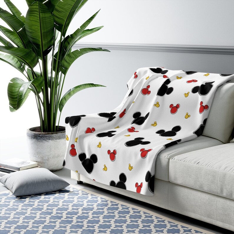 K&ouml;nnte beinhalten: Wei&szlig;e Fleece-Decke mit einem sich wiederholenden Muster aus schwarzen Mickey-Mouse-Ohren und roten und gelben Akzenten.