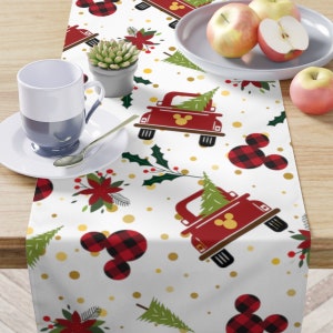 May include: White table runner featuring a red truck carrying a Christmas tree, red and black buffalo check Mickey Mouse ears, holly, poinsettias, and gold polka dots.
