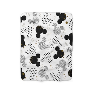 Puede incluir: Manta de forro polar blanca con un patr&oacute;n geom&eacute;trico negro y dorado con siluetas de Mickey Mouse. El patr&oacute;n incluye lunares, rayas y cabezas de Mickey Mouse negras.