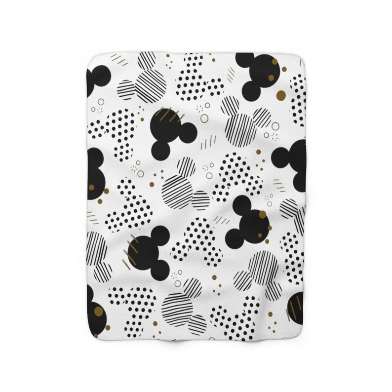 Puede incluir: Manta de forro polar blanca con un patr&oacute;n geom&eacute;trico negro y dorado con siluetas de Mickey Mouse. El patr&oacute;n incluye lunares, rayas y cabezas de Mickey Mouse negras.