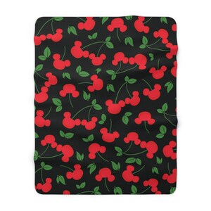 May include: A black and red patterned fleece blanket with a repeating pattern of red cherries with green stems and leaves. The cherries are shaped like Mickey Mouse ears.