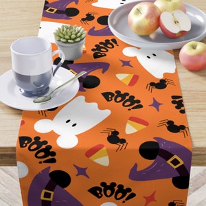 May include: Orange table runner with a Halloween theme. The runner features a repeating pattern of ghosts, witches' hats, candy corn, spiders, and the word "Boo!"