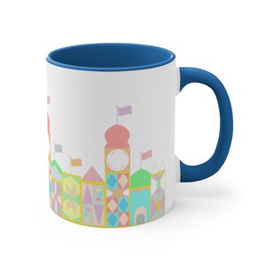 May include: A white ceramic mug with a blue rim and handle. The mug features a colourful illustration of a whimsical building with a clock face and flags.