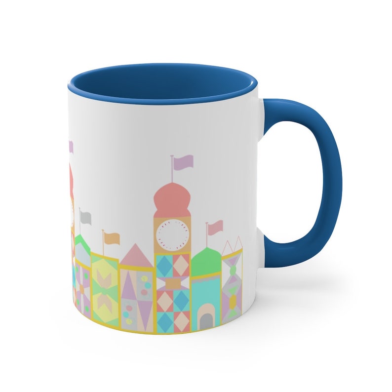 May include: A white ceramic mug with a blue rim and handle. The mug features a colourful illustration of a whimsical building with a clock face and flags.