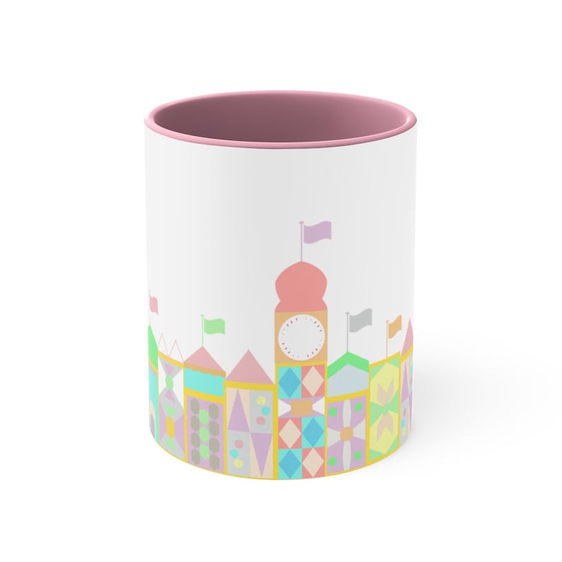 May include: A white ceramic mug with a pink rim features a colourful illustration of a whimsical cityscape with a clock tower and flags. The design is reminiscent of a classic amusement park.