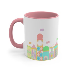 May include: A pink and white ceramic mug featuring a colourful illustration of a whimsical castle with flags and a clock on the front.
