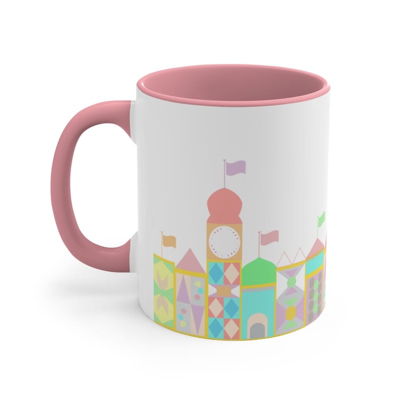 May include: A pink and white ceramic mug featuring a colourful illustration of a whimsical castle with flags and a clock on the front.