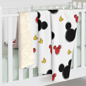 K&ouml;nnte beinhalten: Eine wei&szlig;e Babydecke mit einem sich wiederholenden Muster aus schwarzen Mickey-Maus-K&ouml;pfen und roten und gelben Mickey-Maus-Ohren. Die Decke h&auml;ngt an einem wei&szlig;en Babybett.