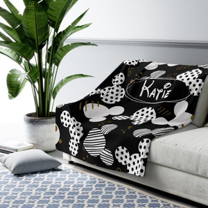Puede incluir: Manta de tiro negra y blanca con un patr&oacute;n de Mickey Mouse y detalles dorados. La manta tiene el nombre "Katie" escrito en ella.