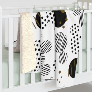 Puede incluir: Una manta de forro polar blanca, negra y dorada con un dise&ntilde;o geom&eacute;trico de c&iacute;rculos y rayas. La manta est&aacute; doblada y drapeada sobre una cuna blanca.