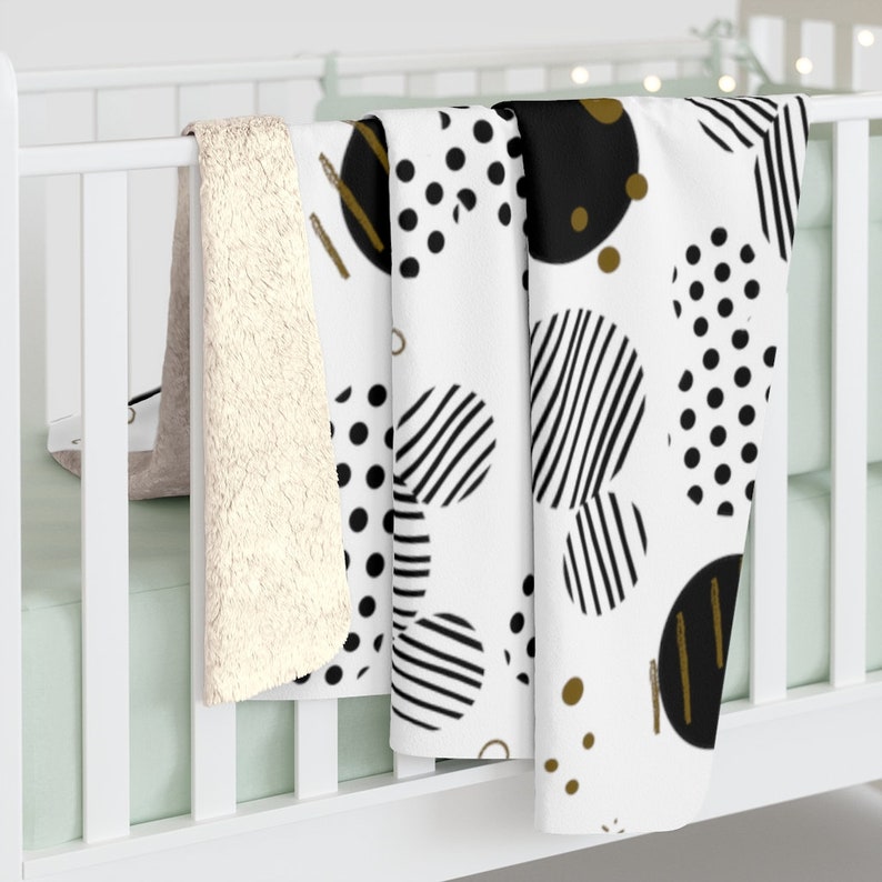 Puede incluir: Una manta de forro polar blanca, negra y dorada con un dise&ntilde;o geom&eacute;trico de c&iacute;rculos y rayas. La manta est&aacute; doblada y drapeada sobre una cuna blanca.