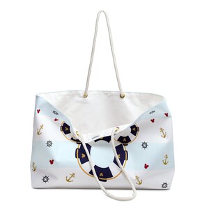K&ouml;nnte beinhalten: Eine wei&szlig;e Canvas-Tragetasche mit blau-wei&szlig;en Streifen und nautischem Thema. Die Tasche zeigt einen Rettungsring mit Mickey-Maus-Ohren, Ankern und Schiffsr&auml;dern. Die Tasche hat wei&szlig;e Seilgriffe.