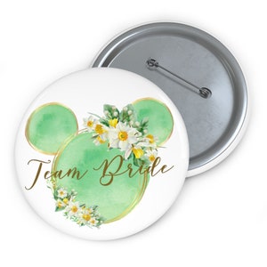 May include: A green and gold button with a floral design and the text "Team Bride".