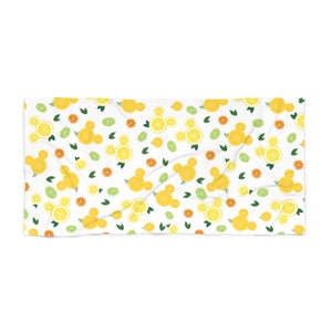 Puede incluir: Una toalla de playa blanca con un patr&oacute;n repetido de rodajas de lim&oacute;n amarillo, hojas verdes y rodajas de c&iacute;tricos naranjas. Las rodajas de lim&oacute;n est&aacute;n dispuestas en forma de cabezas de Mickey Mouse.