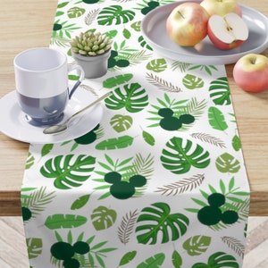 May include: White table runner with a green tropical leaf pattern and Mickey Mouse silhouettes. The runner is on a wooden table with a plate of apples and a mug.
