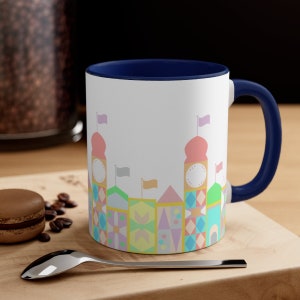 May include: A white ceramic coffee mug with a blue rim and handle. The mug features a colourful illustration of a whimsical castle with pastel pink, yellow, blue, and green colours.