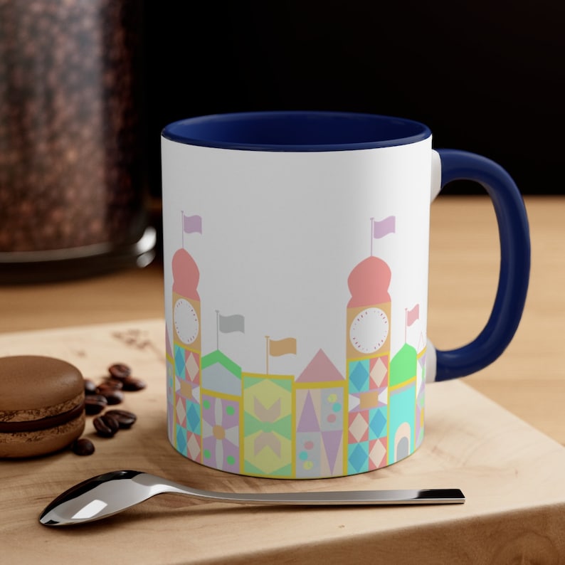 May include: A white ceramic coffee mug with a blue rim and handle. The mug features a colourful illustration of a whimsical castle with pastel pink, yellow, blue, and green colours.