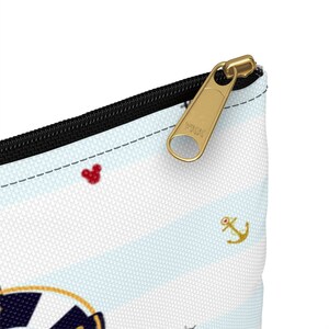 K&ouml;nnte beinhalten: Eine wei&szlig;e Rei&szlig;verschlusstasche mit einem blau-wei&szlig; gestreiften Muster. Die Tasche hat einen goldenen Rei&szlig;verschluss mit dem Text "YKK" darauf. Die Tasche hat auch ein rotes Mickey Mouse-Symbol und ein goldenes Anker-Symbol.