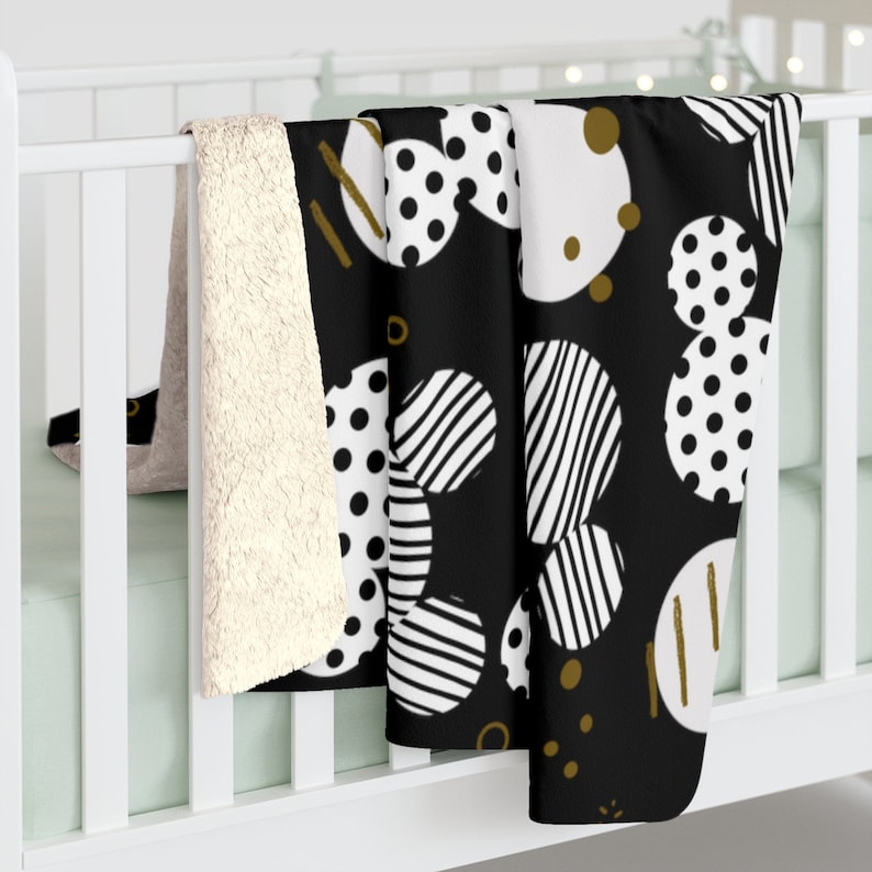 Puede incluir: Manta de tiro de patr&oacute;n blanco y negro con acentos dorados. La manta presenta un patr&oacute;n repetitivo de c&iacute;rculos con lunares, rayas y c&iacute;rculos blancos s&oacute;lidos. La manta est&aacute; drapeada sobre una cuna blanca.