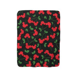 May include: A black and red patterned fleece blanket with a repeating pattern of red cherries with green stems and leaves. The cherries are shaped like Mickey Mouse ears.