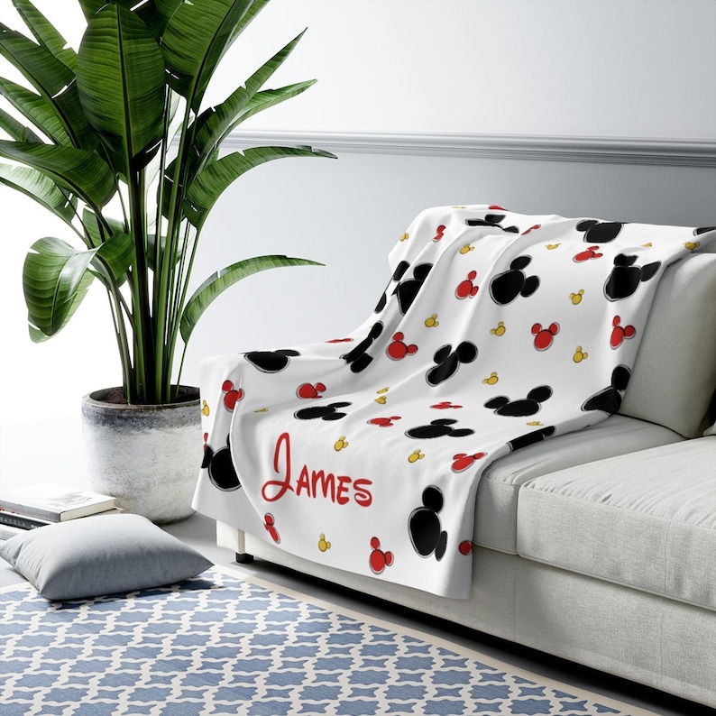 K&ouml;nnte beinhalten: Wei&szlig;e Fleece-Decke mit einem schwarzen und roten Mickey-Mouse-Muster. Die Decke tr&auml;gt den Namen "James" in roter Kursivschrift.