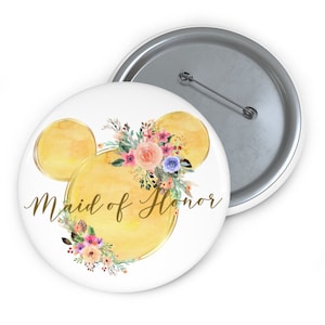May include: A white button with a gold Mickey Mouse head design and the words "Maid of Honor" in gold script. The design includes yellow watercolor circles and a floral arrangement with pink, peach, and blue flowers.