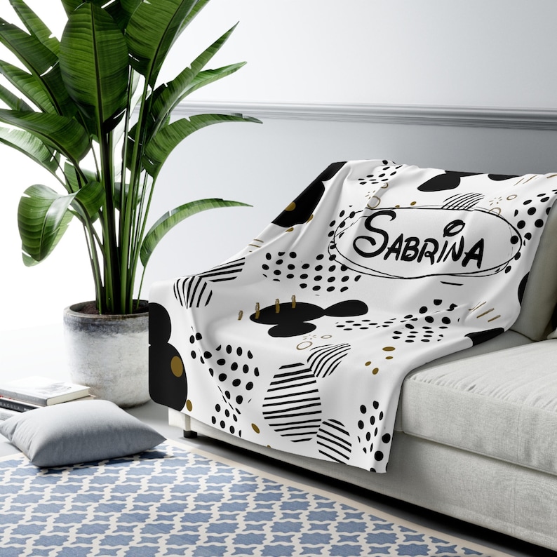 Puede incluir: Manta blanca con un patr&oacute;n abstracto en blanco y negro y detalles dorados. El nombre Sabrina est&aacute; escrito en un &oacute;valo negro en el centro de la manta.