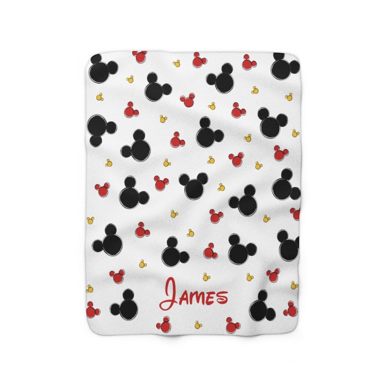 K&ouml;nnte beinhalten: Eine wei&szlig;e Fleece-Decke mit einem schwarzen und roten Mickey-Mouse-Muster. Der Name "James" ist in roter Kursivschrift am unteren Rand der Decke geschrieben.