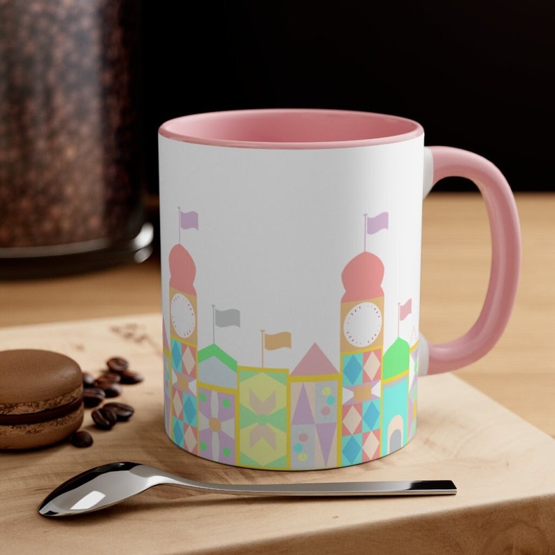 May include: A white ceramic mug with a pink rim and handle. The mug features a colourful illustration of a whimsical cityscape with pastel-coloured buildings and flags.