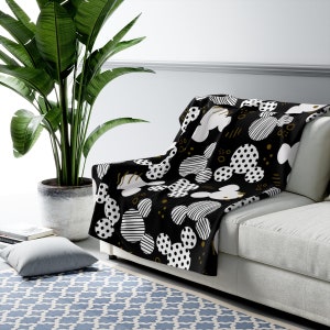 Puede incluir: Manta de tiro negra y blanca con un patr&oacute;n geom&eacute;trico de cabezas de Mickey Mouse. La manta est&aacute; sobre un sof&aacute; blanco con una almohada de patr&oacute;n negro y blanco.