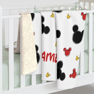 K&ouml;nnte beinhalten: Wei&szlig;e Babydecke mit Mickey Mouse-Motiv. Die Decke zeigt schwarze und rote Mickey Mouse-K&ouml;pfe und gelbe Kreise. Der Name "Ama" ist in Rot auf der Decke geschrieben.