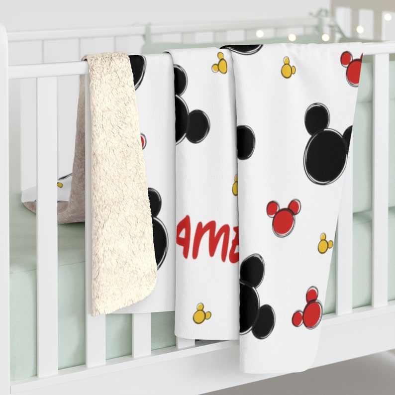 K&ouml;nnte beinhalten: Wei&szlig;e Babydecke mit Mickey Mouse-Motiv. Die Decke zeigt schwarze und rote Mickey Mouse-K&ouml;pfe und gelbe Kreise. Der Name "Ama" ist in Rot auf der Decke geschrieben.