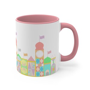 May include: A white ceramic mug with a pink rim and handle. The mug features a colourful illustration of a whimsical building with a clock tower and flags.
