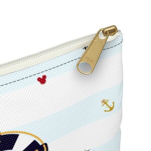 K&ouml;nnte beinhalten: Eine wei&szlig;e Rei&szlig;verschlusstasche mit einem blau-wei&szlig; gestreiften Muster. Die Tasche hat einen goldenen Rei&szlig;verschluss mit dem Text "YKK" darauf. Die Tasche hat auch ein rotes Mickey Mouse-Symbol und ein goldenes Anker-Symbol.