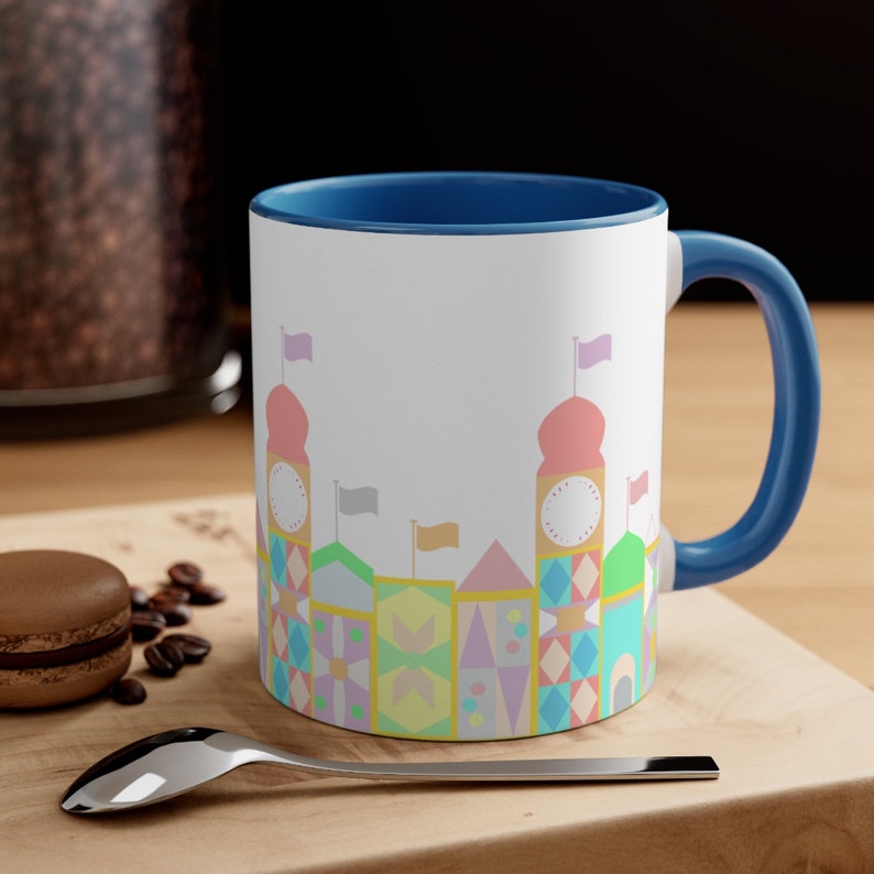 May include: A white ceramic coffee mug with a blue rim and handle. The mug features a colourful illustration of a whimsical cityscape with pastel-coloured buildings and flags.
