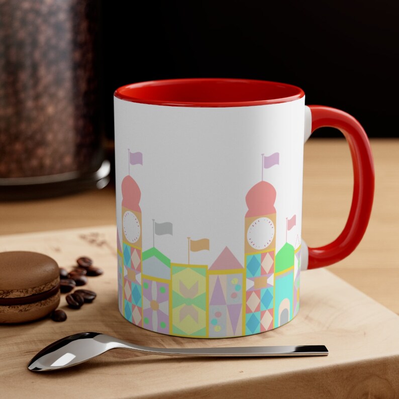 May include: A white ceramic mug with a red rim and handle. The mug features a colourful illustration of a whimsical cityscape with pastel-coloured buildings and flags.