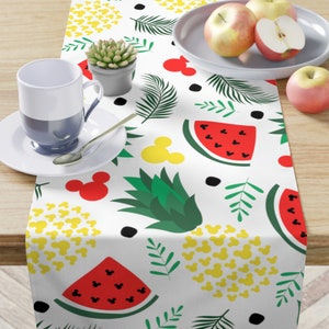 May include: White table runner with a repeating pattern of red watermelon slices, green pineapple, yellow Mickey Mouse heads, and green leaves.