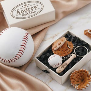 Puede incluir: Una pelota de béisbol blanca con costuras rojas y una caja de regalo con el nombre "Andrew". También hay llaveros con temática de béisbol con una pelota, un guante y un bate. Los llaveros están en una caja de regalo con relleno negro.