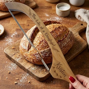 Op de afbeelding: Een houten broodmes met een roestvrijstalen mes snijdt een brood op een houten snijplank. Op het broodmes staat de tekst "The Life of a Doughgirl" en de naam "Kristen" gegraveerd.