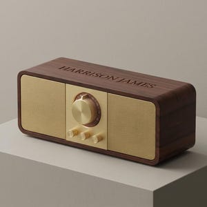 Peut inclure: Une radio rectangulaire de style rétro avec une finition en bois foncé. La radio présente une façade de haut-parleur et des boutons de commande dorés. Le texte "HARRISON JAMES" est gravé sur le dessus.